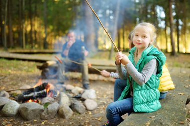 Sevimli kız kardeşler ve babaları şenlik ateşinde sopalarla marshmallow kızartıyorlar. Kamp ateşinde eğlenen çocuklar. Sonbahar ormanında çocuklarla kamp.