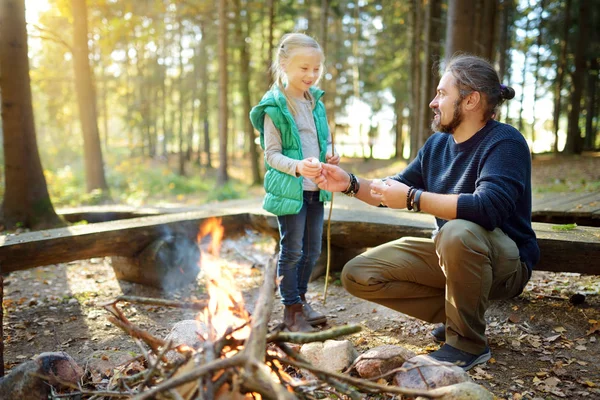 Aranyos fiatal lány, a tanulás kezdeni a tábortűz. Apja tanítja a lányát, hogy a tüzet. Gyermek szórakozik a tábortűz. Camping gyerekekkel őszi erdő. — Stock Fotó