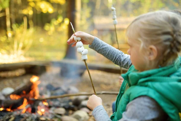 Adorable jovencita asando malvaviscos en el palo en la hoguera. Un niño divirtiéndose en el campamento. Camping con niños en bosque otoñal . — Foto de Stock
