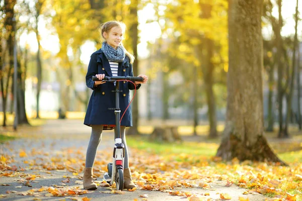 Adorable young girl riding her scooter in a city park on sunny autumn evening. Pretty preteen child riding a roller. — Stock Photo, Image