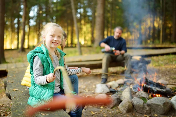 Sevimli kız kardeşler ve babaları şenlik ateşinde sopalarla marshmallow kızartıyorlar. Kamp ateşinde eğlenen çocuklar. Sonbahar ormanında çocuklarla kamp.