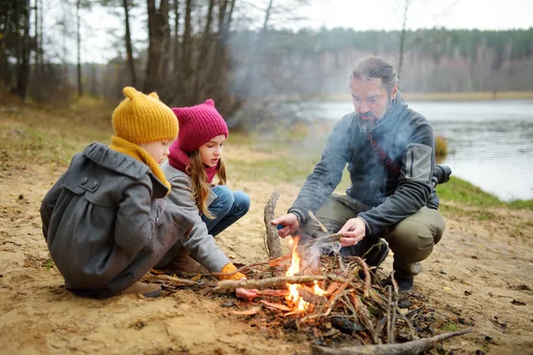 Niedliche kleine Schwestern und ihr Vater sitzen an einem kalten Herbsttag am Lagerfeuer. Kinder haben Spaß am Lagerfeuer. Zelten mit Kindern im Herbstwald. — Stockfoto