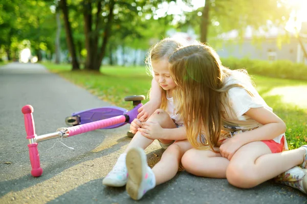 Schattig meisje trooting haar zusje nadat ze viel van haar scooter in zomerpark. Kind krijgen gewond tijdens het rijden een kick scooter. — Stockfoto