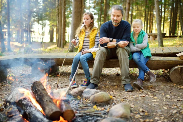 Niedliche kleine Schwestern und ihr Vater braten Marshmallows am Lagerfeuer. Kinder haben Spaß am Lagerfeuer. Zelten mit Kindern im Herbstwald. — Stockfoto