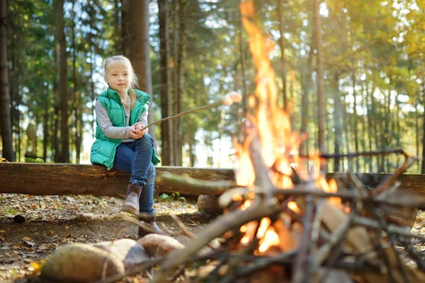 Imádnivaló fiatal lány pörkölés mályvacukrot a botot máglyán. Gyermek szórakozik a tábortűz. Kemping gyerekekkel ősszel erdőben. — Stock Fotó