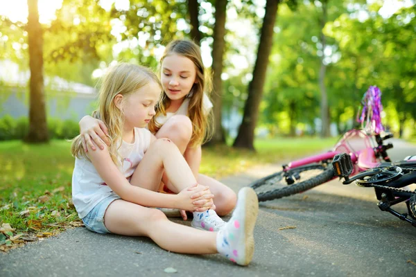 Schattig meisje trooting haar zusje nadat ze viel van haar fiets in zomerpark. Kind dat gewond raakt tijdens het rijden op een fiets. — Stockfoto