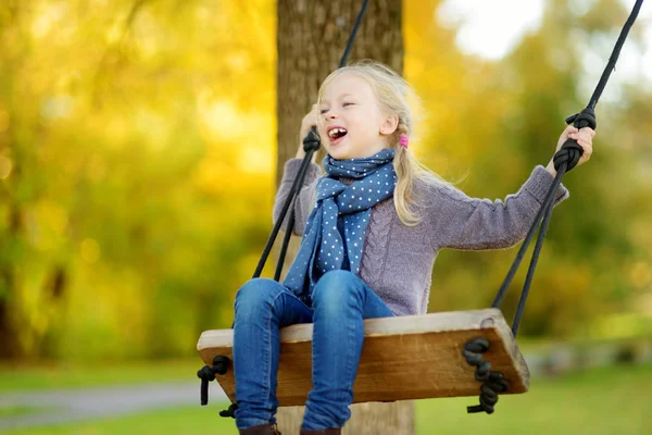 Gadis muda yang lucu bersenang-senang di ayunan di taman musim gugur yang cerah. Akhir pekan keluarga di kota . — Stok Foto
