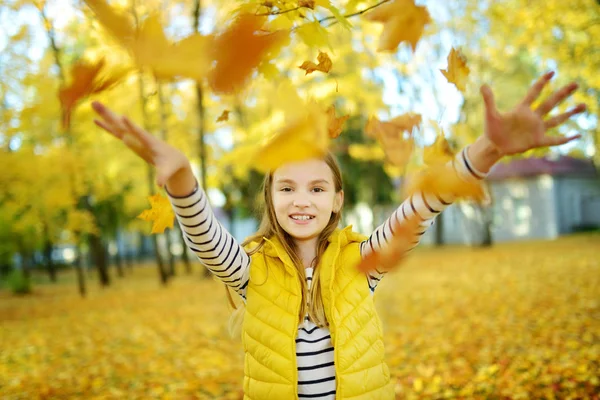 Adorable chica joven divirtiéndose en hermoso día de otoño. Feliz niño jugando en el parque de otoño. Niño recogiendo follaje de otoño amarillo . — Foto de Stock