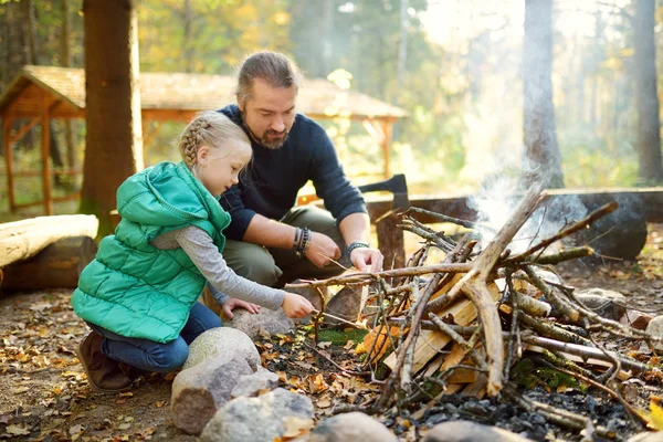 Nettes junges Mädchen, das lernt, ein Lagerfeuer zu entzünden. Der Vater lehrt seine Tochter, ein Feuer zu machen. Kinder amüsieren sich am Lagerfeuer. Zelten mit Kindern im Herbstwald. — Stockfoto