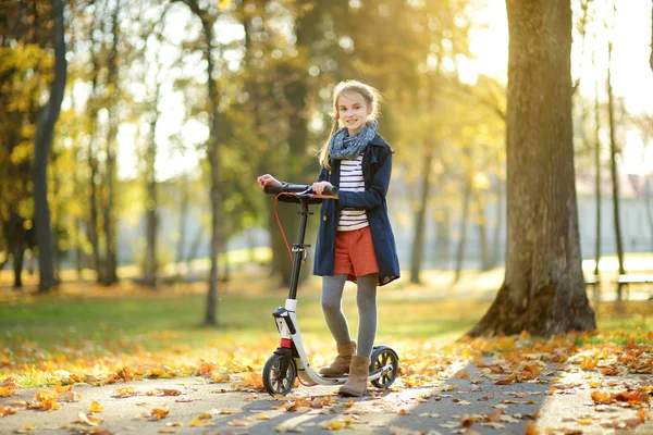 Menina adorável montando sua scooter em um parque da cidade na noite de outono ensolarada. Bela criança pré-adolescente montando um rolo . — Fotografia de Stock