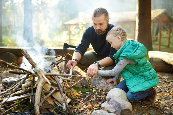 Menina bonito aprender a iniciar uma fogueira. O pai a ensinar a filha a fazer uma fogueira. Criança a divertir-se no acampamento. Acampar com crianças na floresta de outono . — Fotografia de Stock