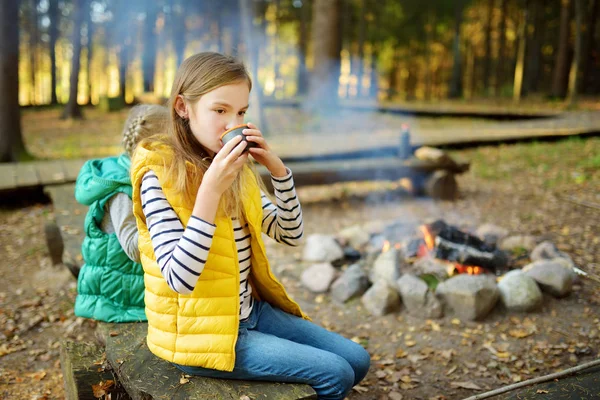 Linda chica joven bebiendo té y tostando malvaviscos en palo en la hoguera. Un niño divirtiéndose en el campamento. Camping con niños en bosque otoñal . — Foto de Stock