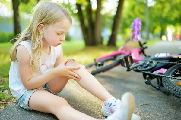 Schattig klein meisje zittend op de grond na vallen van haar fiets in zomerpark. Kind dat gewond raakt tijdens het rijden op een fiets. — Stockfoto