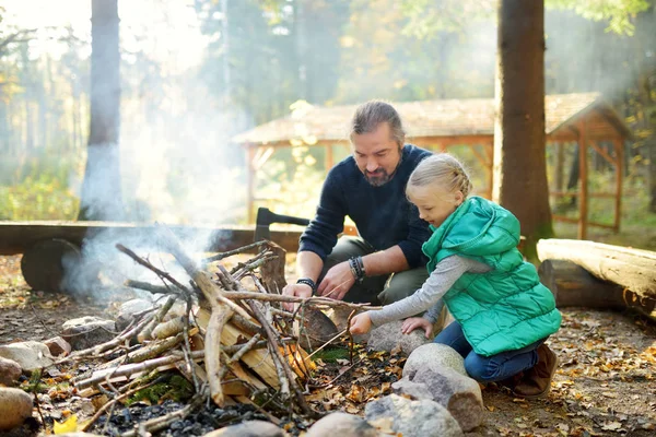 Cute young girl learning to start a bonfire. Father teaching her daughter to make a fire. Child having fun at camp fire. Camping with kids in fall forest. — Stock Photo, Image