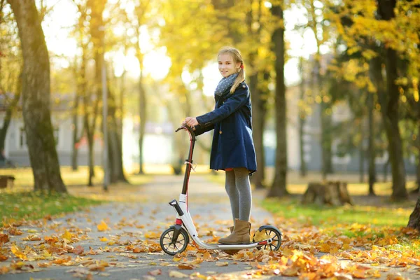 Adorabile ragazza che cavalca il suo scooter in un parco cittadino la sera d'autunno soleggiata. Abbastanza bambino preadolescente cavalcando un rullo . — Foto Stock