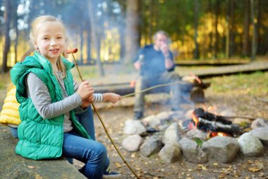 Sevimli kız kardeşler ve babaları şenlik ateşinde sopalarla marshmallow kızartıyorlar. Kamp ateşinde eğlenen çocuklar. Sonbahar ormanında çocuklarla kamp.