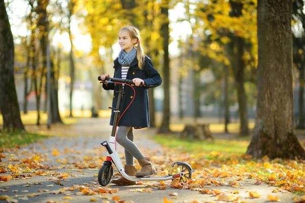 Adorabile ragazza che cavalca il suo scooter in un parco cittadino la sera d'autunno soleggiata. Abbastanza bambino preadolescente cavalcando un rullo . — Foto Stock