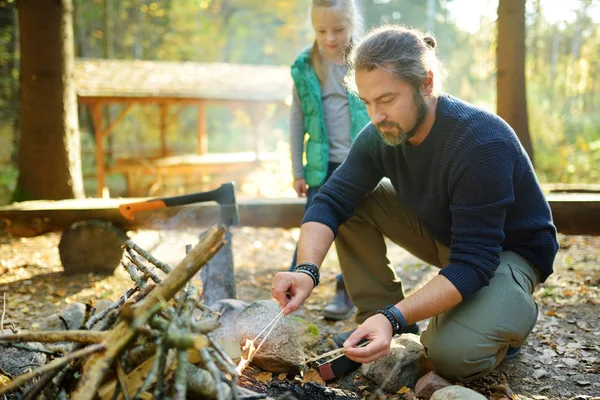 Nettes junges Mädchen, das lernt, ein Lagerfeuer zu entzünden. Der Vater lehrt seine Tochter, ein Feuer zu machen. Kinder amüsieren sich am Lagerfeuer. Zelten mit Kindern im Herbstwald. — Stockfoto