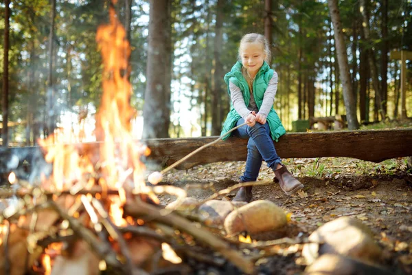 Adorable jovencita asando malvaviscos en el palo en la hoguera. Un niño divirtiéndose en el campamento. Camping con niños en bosque otoñal . — Foto de Stock