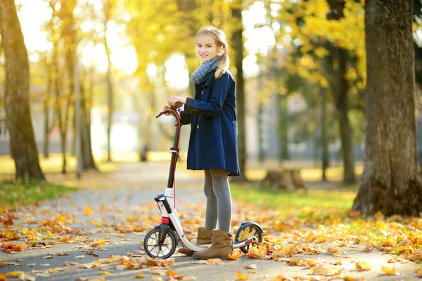 Schattig jong meisje rijden haar scooter in een stadspark op zonnige herfstavond. Mooie Preteen kind rijden een roller. — Stockfoto