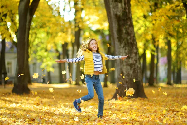 Adorable chica joven divirtiéndose en hermoso día de otoño. Feliz niño jugando en el parque de otoño. Niño recogiendo follaje de otoño amarillo . — Foto de Stock