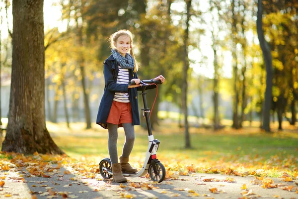 Adorable young girl riding her scooter in a city park on sunny autumn evening. Pretty preteen child riding a roller. — Stock Photo, Image
