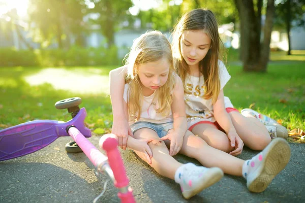 Schattig meisje trooting haar zusje nadat ze viel van haar scooter in zomerpark. Kind krijgen gewond tijdens het rijden een kick scooter. — Stockfoto