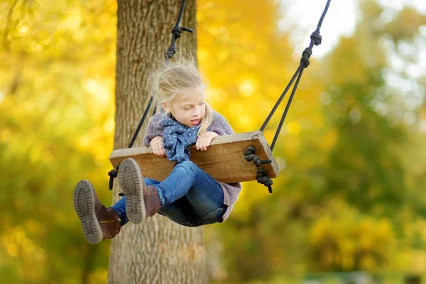 Roztomilá mladá dívka se baví na houpačce ve slunném podzimním parku. Rodinný víkend ve městě. — Stock fotografie