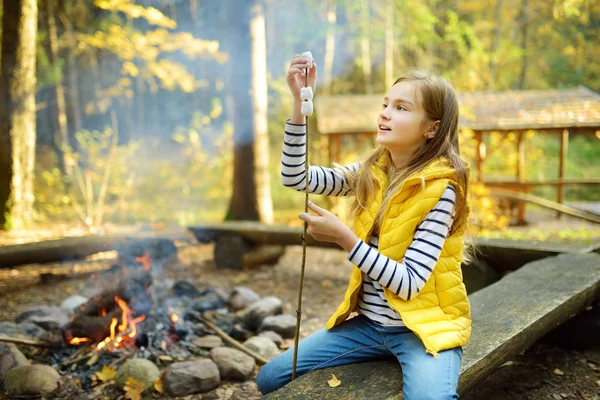 Urocza młoda dziewczyna prażenie marshmallows na kij na ognisko. Dziecko bawią się ogniem obozu. Kemping z dziećmi w lesie jesienne. — Zdjęcie stockowe