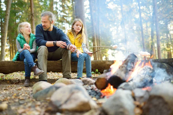 Niedliche kleine Schwestern und ihr Vater braten Marshmallows am Lagerfeuer. Kinder haben Spaß am Lagerfeuer. Zelten mit Kindern im Herbstwald. — Stockfoto