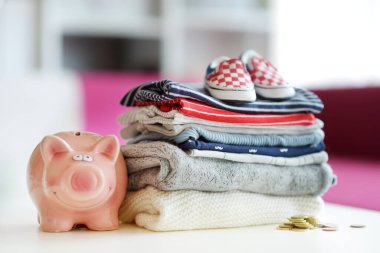 Bir yığın bebek kıyafeti, küçük ayakkabılar ve kumbara. Ebeveyn masrafları konsepti. Bebek bütçesi hazırlıyorum. Yeni doğmuş bir bebek için para biriktiriyorum. Yeni bir bebek için bütçe ayırıyorum..