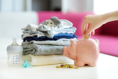 Bir yığın bebek giysisi, kumbara, emzik ve beslenme şişesi. Ebeveyn masrafları konsepti. Bebek bütçesi hazırlıyorum. Yeni doğmuş bir bebek için para biriktiriyorum. Yeni bir bebek için bütçe ayırıyorum..