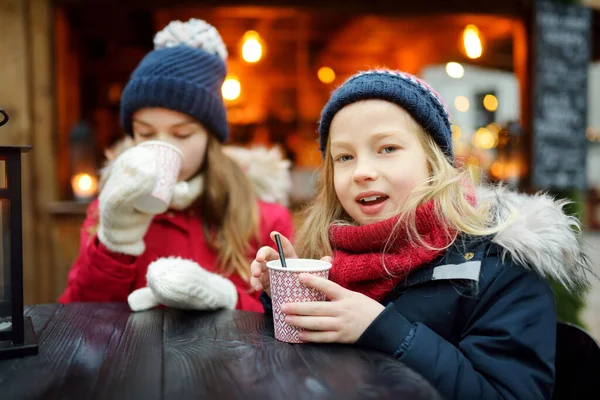 ラトビアのリガで伝統的なクリスマスフェアでホットチョコレートを飲む2人の愛らしい姉妹 クリスマスマーケットでお菓子やキャンディー ジンジャーブレッドを楽しむ子供たち 家族や子供と冬の時間 — ストック写真