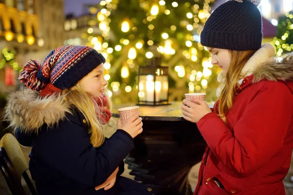 ラトビアのリガで伝統的なクリスマスフェアでホットチョコレートを飲む2人の愛らしい姉妹 クリスマスマーケットでお菓子やキャンディー ジンジャーブレッドを楽しむ子供たち 家族や子供と冬の時間 — ストック写真