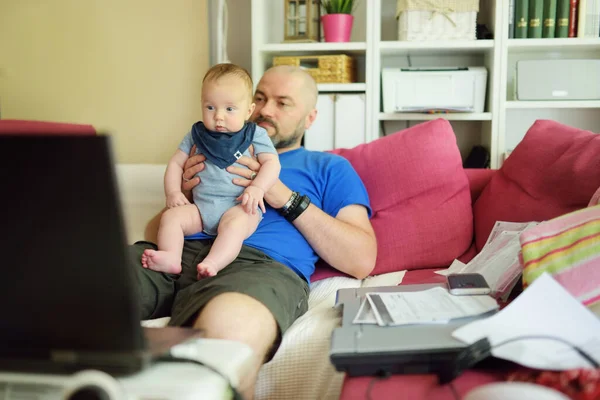年轻的爸爸和孩子在家里的办公室里远程工作 自由职业者在使用笔记本电脑时抱着他的婴儿 客厅里的工作场所检疫期间工作 待在家里 — 图库照片