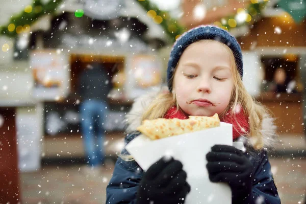 リトアニアのヴィリニュスで伝統的なクリスマスフェアでクレープパンケーキを食べるかわいい女の子 クリスマスマーケットでお菓子やキャンディー ジンジャーブレッドを楽しむ子供たち 家族や子供と冬の時間 — ストック写真