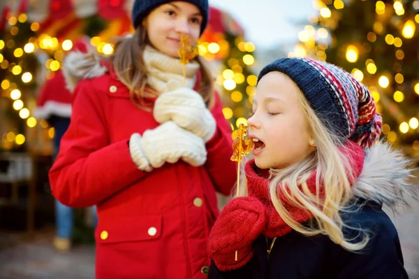 ラトビアのリガで開催される伝統的なクリスマスフェアには 鶏の形をしたロリポップを持つ2人の愛らしい姉妹 クリスマスマーケットでお菓子やキャンディー ジンジャーブレッドを楽しむ子供たち 家族や子供と冬の時間 — ストック写真