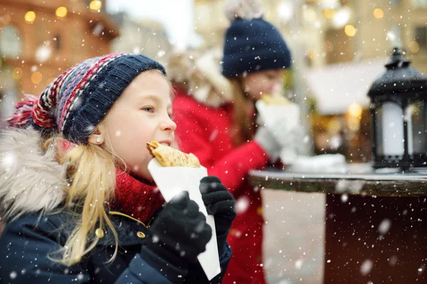 リトアニアのヴィリニュスで伝統的なクリスマスフェアでクレープパンケーキを食べるかわいい若い女の子 クリスマスマーケットでお菓子やキャンディー ジンジャーブレッドを楽しむ子供たち 家族や子供と冬の時間 — ストック写真