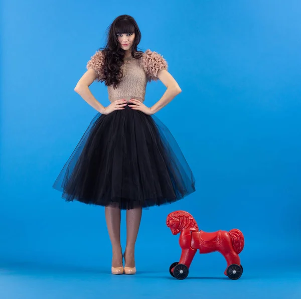 黒のスカートと青色の背景に赤いおもちゃの馬で軽薄なポーズで美しい若いおしゃれな女性 — ストック写真