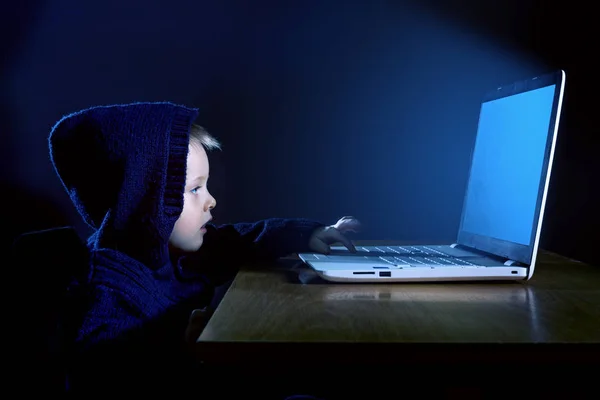 一个穿着黑色头套的小男孩在黑暗中使用笔记本电脑 — 图库照片