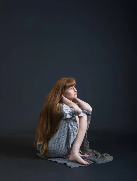 A red-haired young sad woman sitting on floor isolated on a dark background.