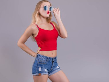 Denim şort ve açık gri arka plan üzerinde kırmızı üst güneş gözlüğü ile şaşırttı genç kadın. Yaz Kadın modası.