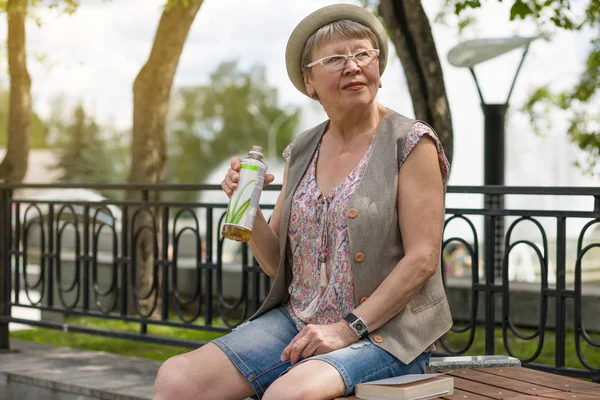 快乐老妇人拿着一瓶饮料 坐在夏天公园的长椅上 — 图库照片
