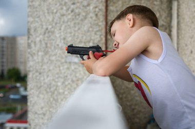 Küçük bir çocuk, çok katlı bir binanın kütüğünde duran oyuncak bir silahla ateş ediyor. Çocuklarda saldırganlığın dışavurumu.