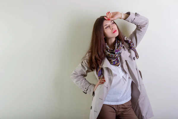 A beautiful fashionable woman in a light raincoat and a neckerchief poses on a light background.