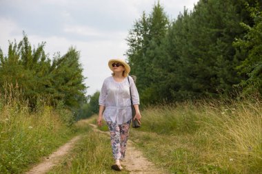 Yaşlı bir kadın kırsal bir yolda yürüyor, yaz doğasının tadını çıkarıyor. Yaşlılar için aktif yaşam tarzı kavramı.
