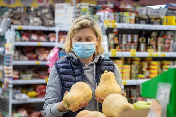 An elderly woman in a medical mask selects vegetables in a grocery store.