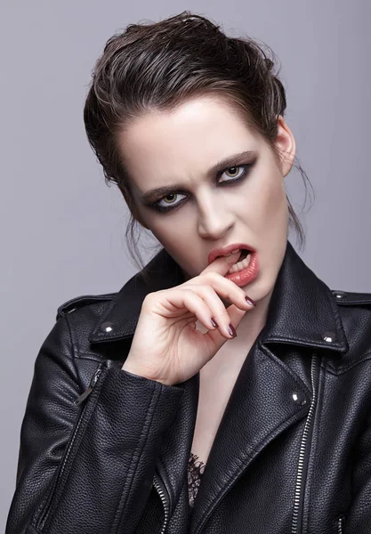 Portrait of female in black leather jacket bites finger. Woman with unusual beauty evening makeup. Girl with perfect skin, green pistachio colour eyes and violet - black shadows make-up.