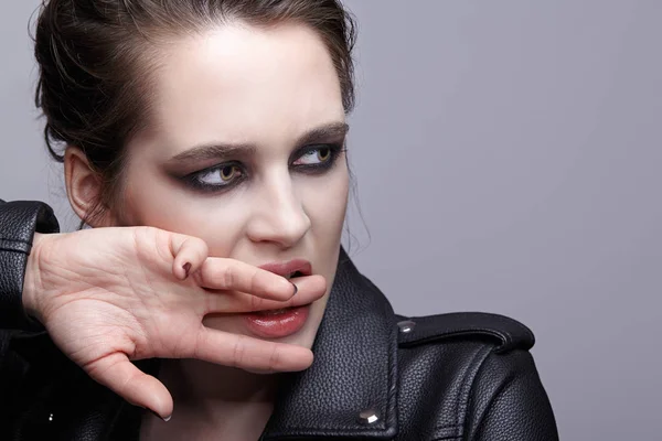 Portrait of female in black leather jacket bites finger. Woman with unusual beauty evening makeup. Girl with perfect skin, green pistachio colour eyes and violet - black shadows make-up.