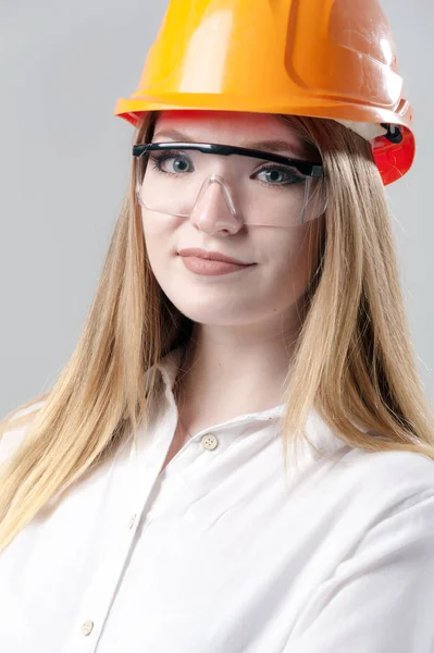 Portrait of a young attractive woman with blond hair in orange helmet on a neutral gray background. — Stock Photo, Image